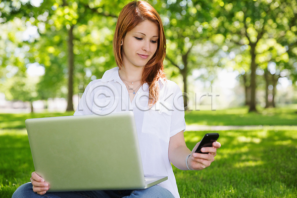 20대 백인 성인 성인여자한명만 여자 청년 한명 JPG 아웃포커스 앞모습 포토 해외이미지 공원 노트북 들기 미소(표정) 상반신 스마트폰 앉기 야외 응시 잔디 해외202007 햇빛